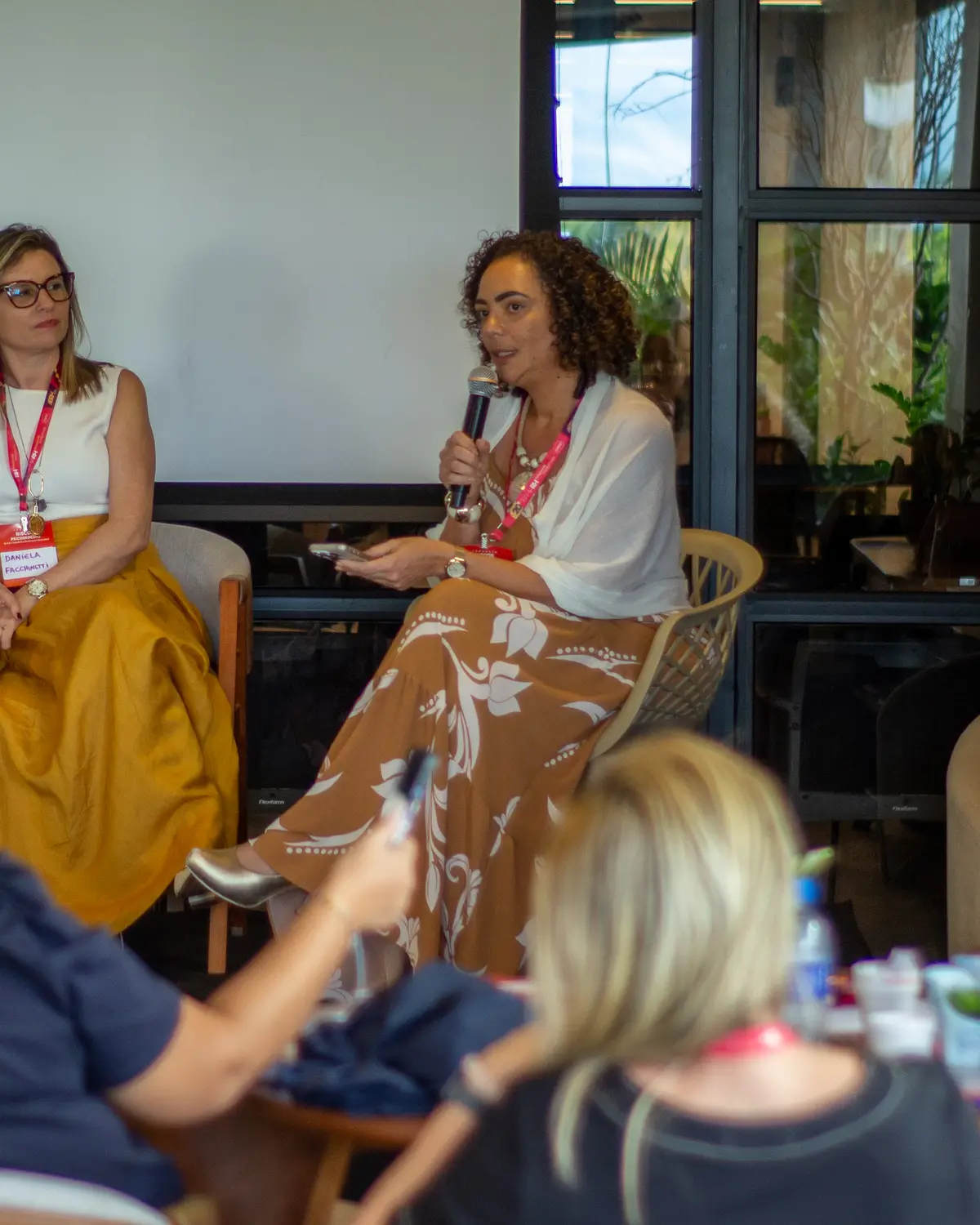 Dra. Ana Paula Teixeira com o microfone em roda de conversa no formato painel, diante de participantes sentados em mesas